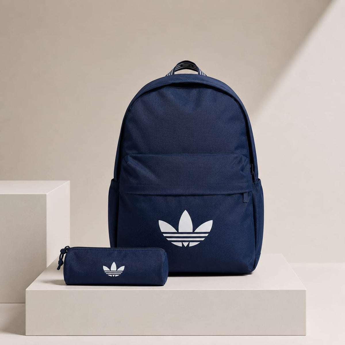 A navy blue Adidas backpack with a white trefoil logo, displayed next to a matching navy blue pencil case on white display blocks against a beige background.