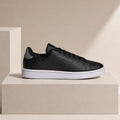 A black leather Adidas Campus sneaker with a white rubber sole, displayed on a beige geometric pedestal against a matching beige background.