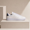 A white Adidas Campus sneaker with a navy blue heel tab, displayed on a minimalist beige pedestal against a matching beige background.