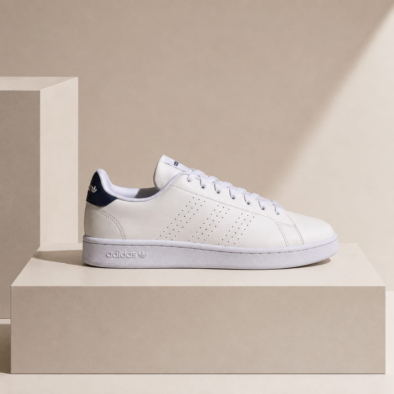 A white Adidas Campus sneaker with a navy blue heel tab, displayed on a minimalist beige pedestal against a matching beige background.
