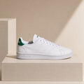 A white Adidas Campus Classic sneaker with a green heel tab, displayed on a beige pedestal against a beige background.