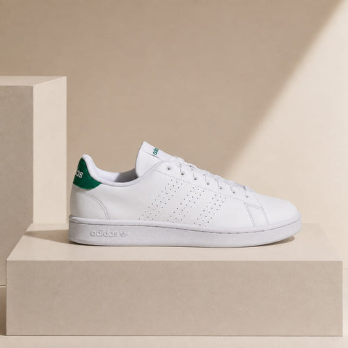 A white Adidas Campus Classic sneaker with a green heel tab, displayed on a beige pedestal against a beige background.