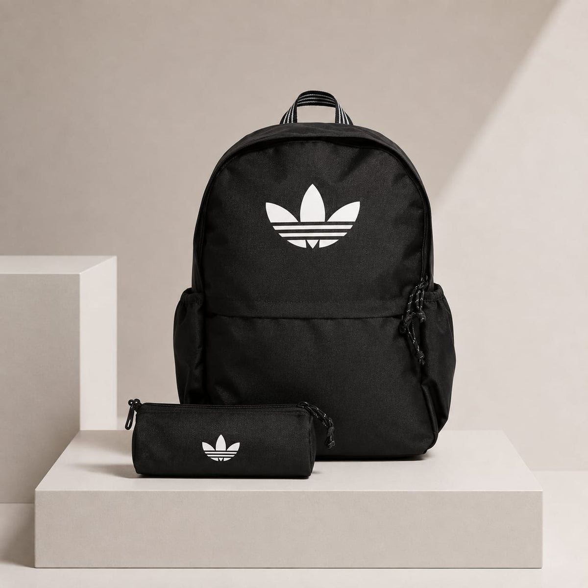 A black Adidas backpack with a white trefoil logo, displayed on a white pedestal next to a matching black pencil case with the same logo, against a beige background.