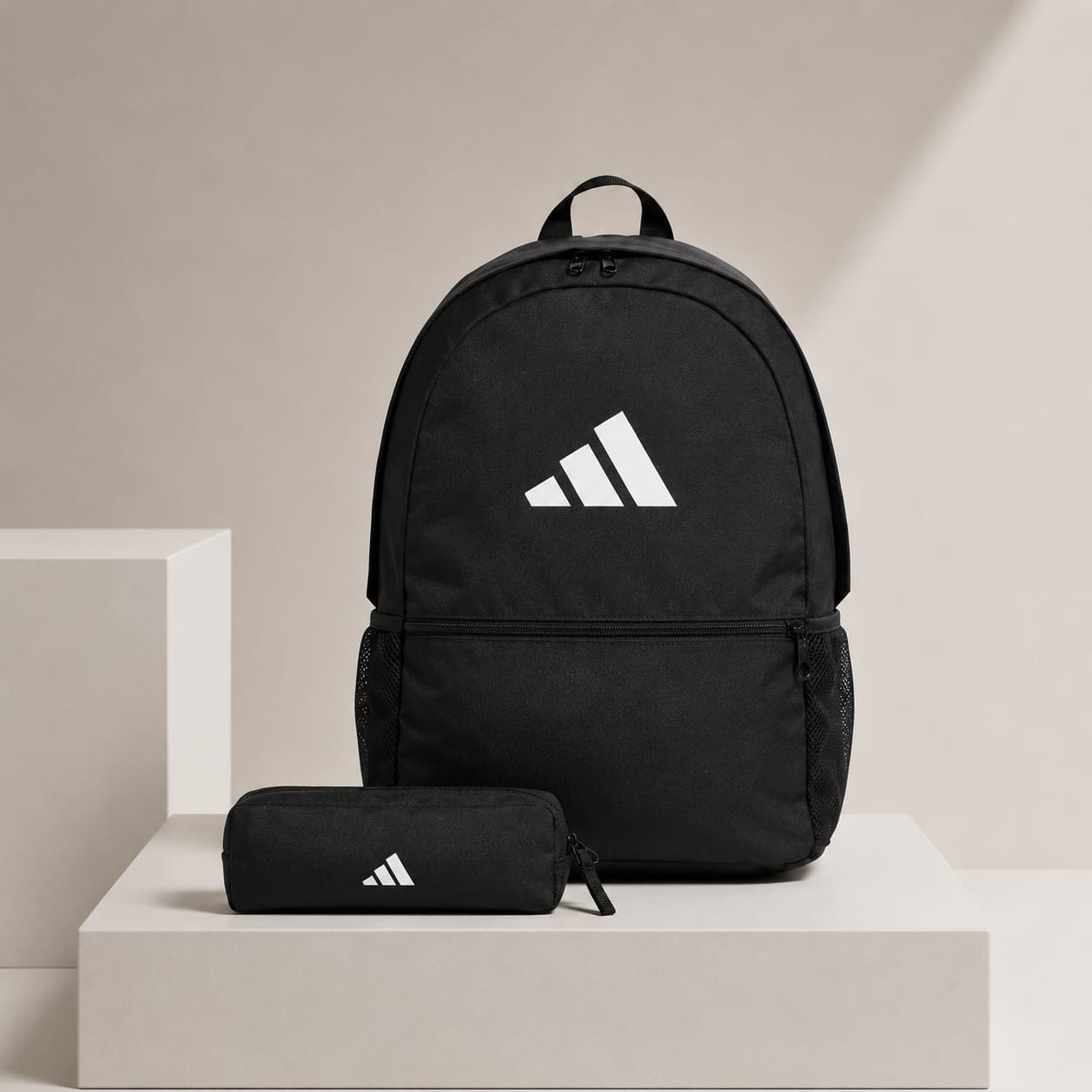 A black Adidas backpack with a white logo is displayed on a light beige pedestal alongside a matching black zippered pouch, set against a minimalist beige background.