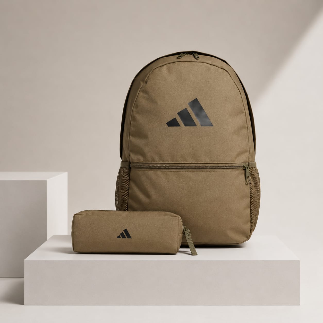 An Adidas backpack in a warm khaki-tan color stands on a white display pedestal next to a matching zippered pouch. Both items feature the black Adidas logo on the front.