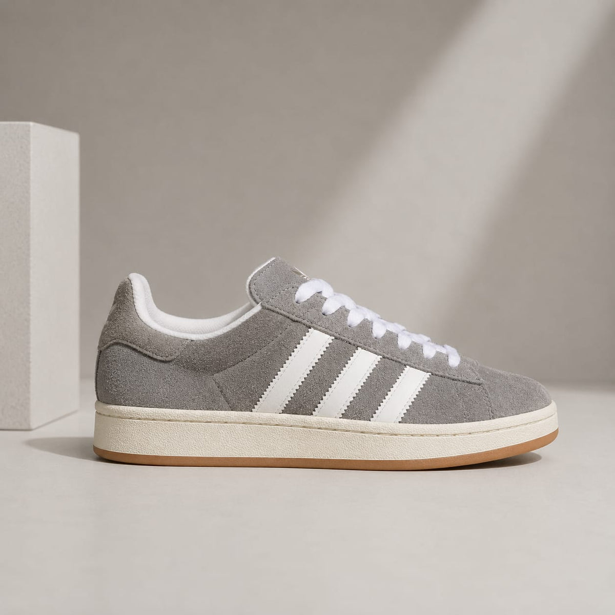 A classic low-top sneaker in gray suede with three white stripes, white laces, and a cream midsole with a gum outsole, shown in profile against a minimalist beige background.