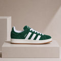 A green suede sneaker with white stripes and white laces, displayed on a beige pedestal against a beige background.