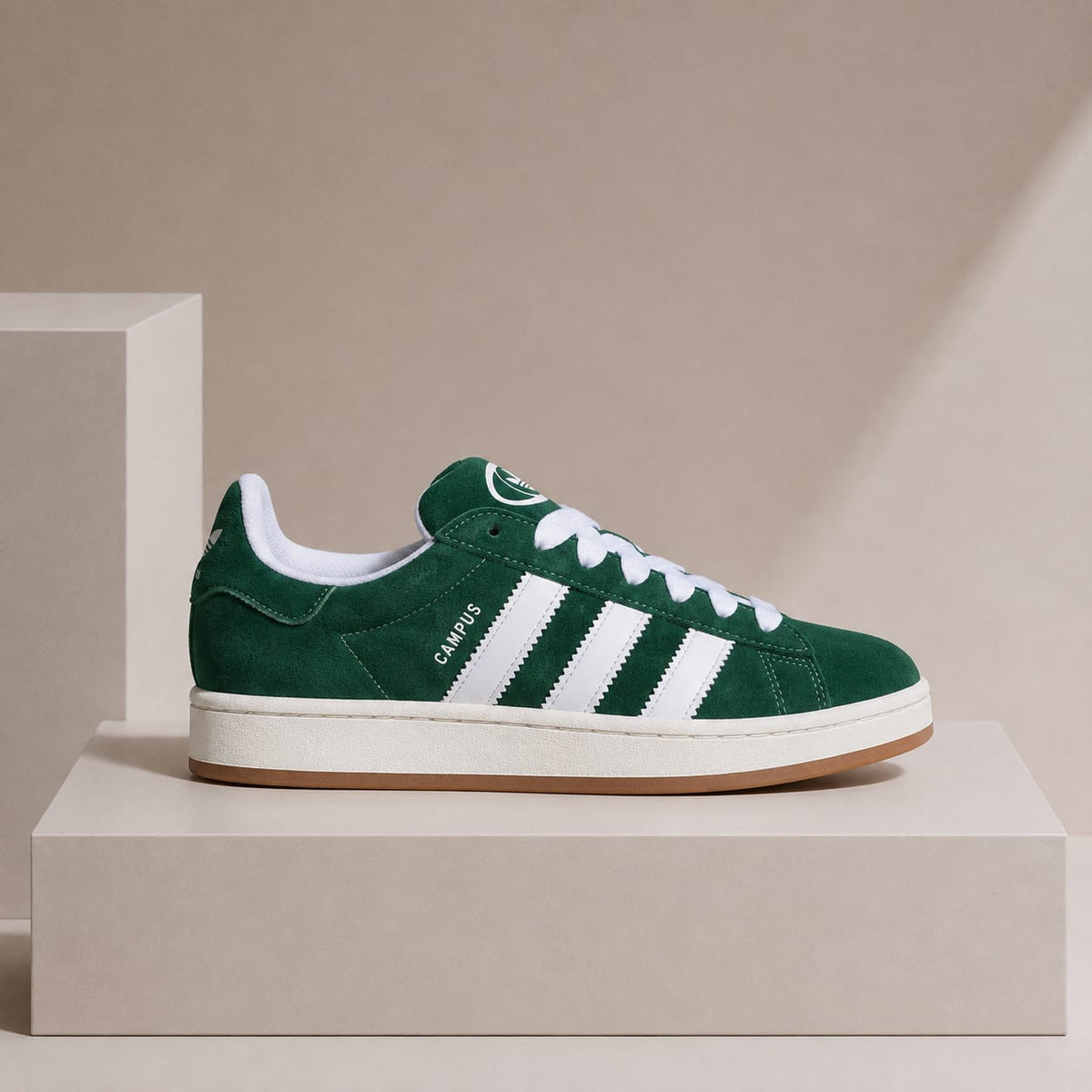 A green suede sneaker with white stripes and white laces, displayed on a beige pedestal against a beige background.