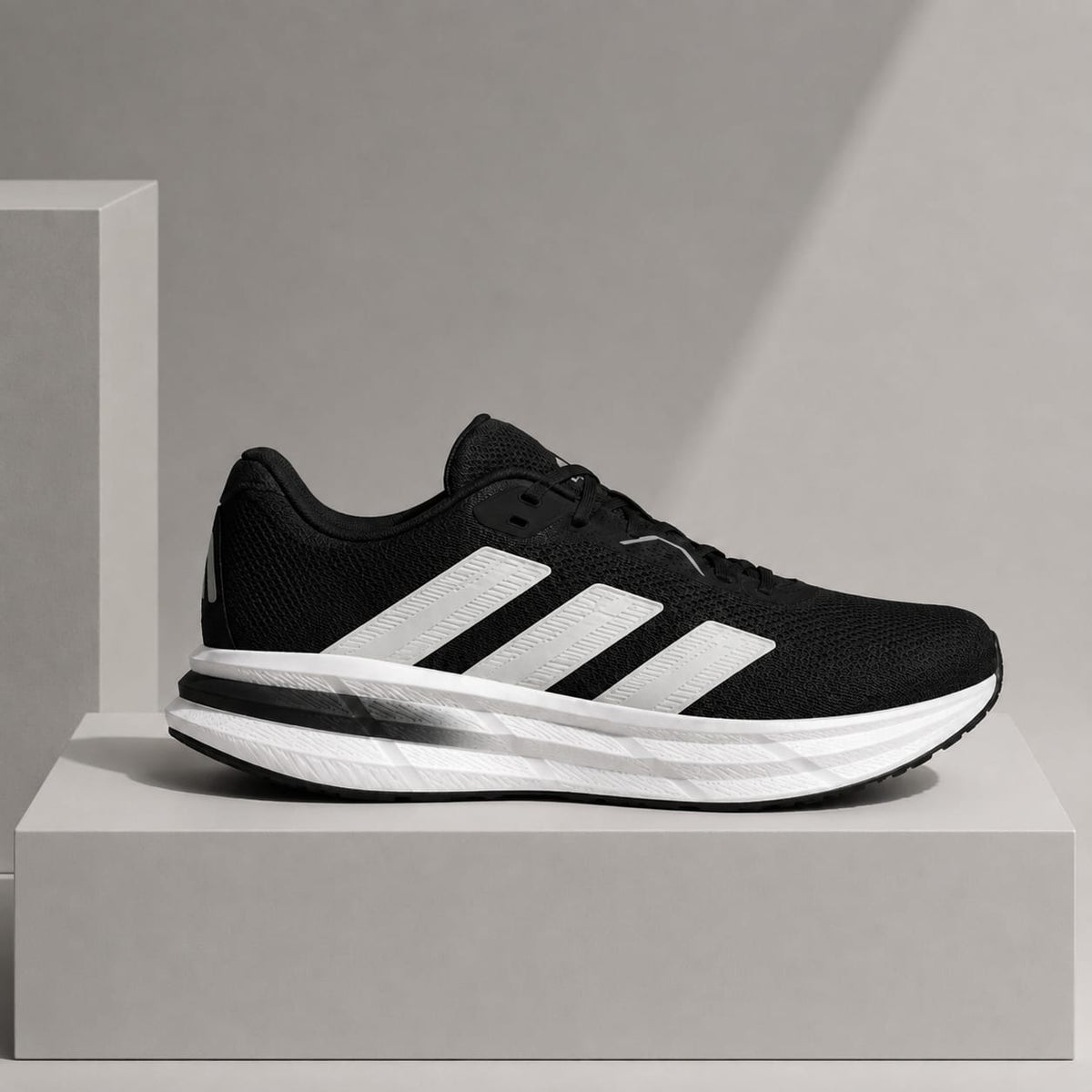 A black and white Adidas running shoe displayed on a minimalist gray pedestal against a neutral gray background.