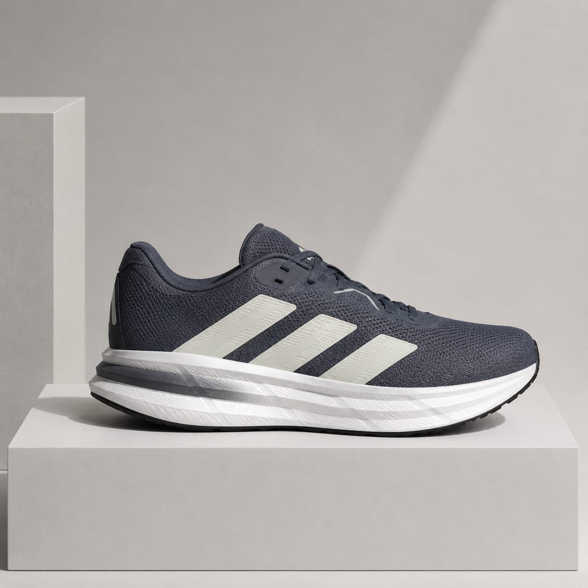 A single Adidas running shoe in navy blue and white, displayed in profile on a light gray pedestal against a neutral gray background.