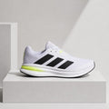 A white Adidas running shoe with black three-stripes and a bright neon yellow-green midsole accent, displayed on a white pedestal against a light gray background.