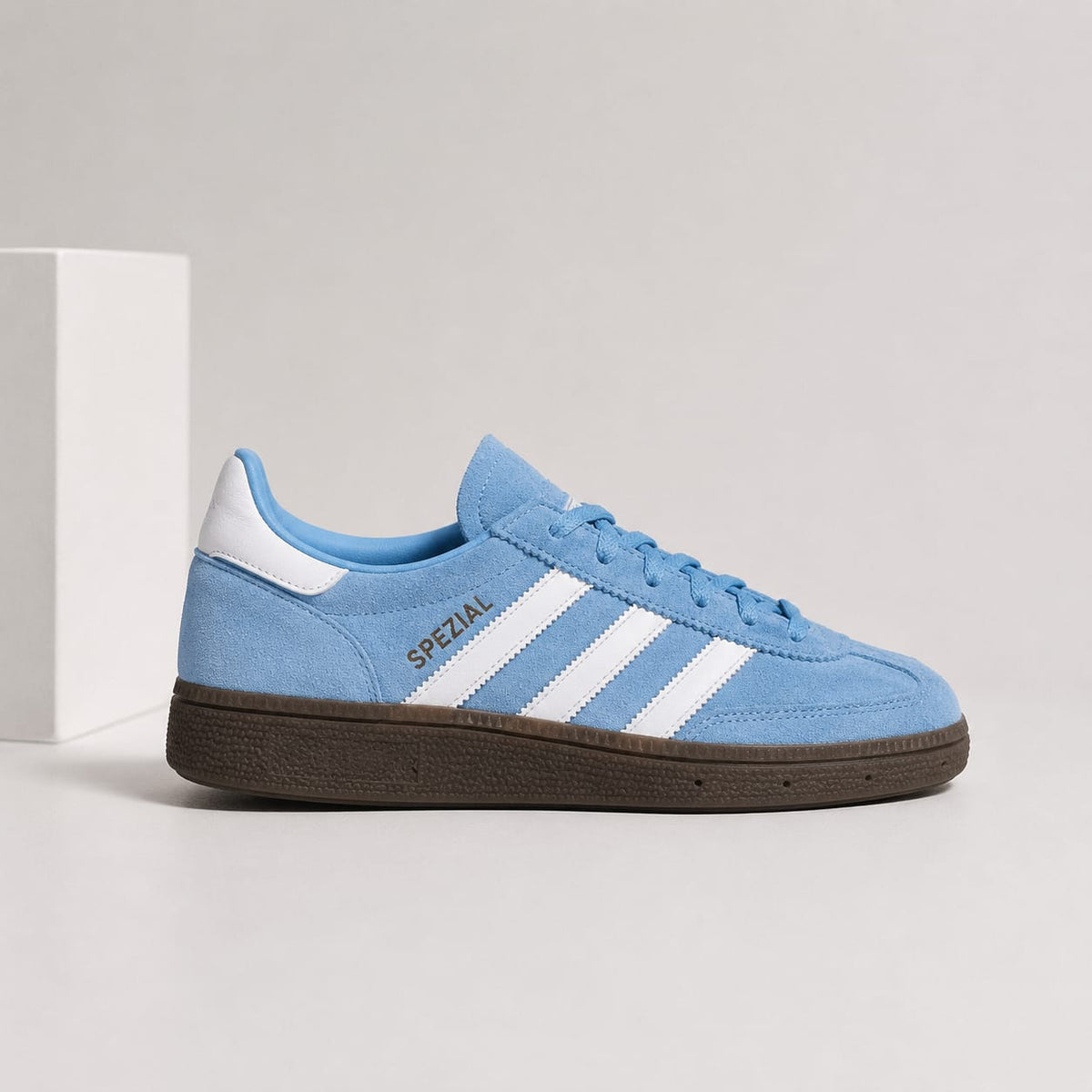 A light blue suede Adidas Gazelle sneaker with white three-stripes, a brown gum sole, and gold 'SPEZIAL' text on the side, shown next to a white block on a light gray background.