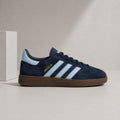 A navy blue suede Adidas Gazelle sneaker with light blue three stripes, a gold 'SPECIALY' logo, and a brown gum sole, displayed on a neutral beige surface next to a white block.