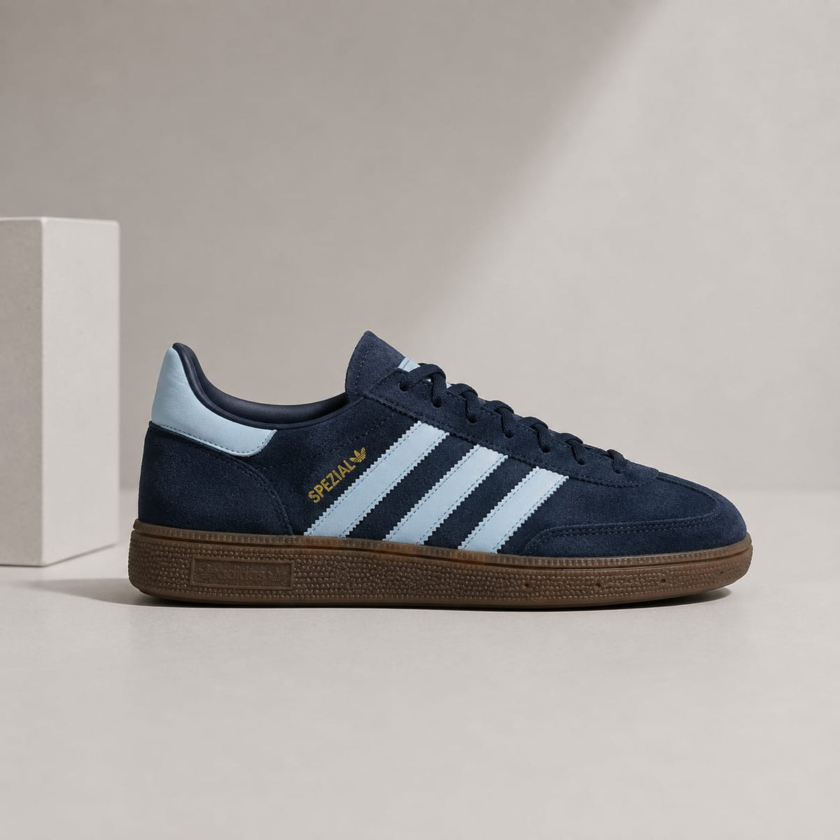 A navy blue suede Adidas Gazelle sneaker with light blue three stripes, a gold 'SPECIALY' logo, and a brown gum sole, displayed on a neutral beige surface next to a white block.