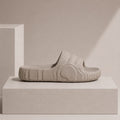 A single light beige slide sandal with a thick, textured wavy sole, displayed on a minimalist beige pedestal against a matching background.