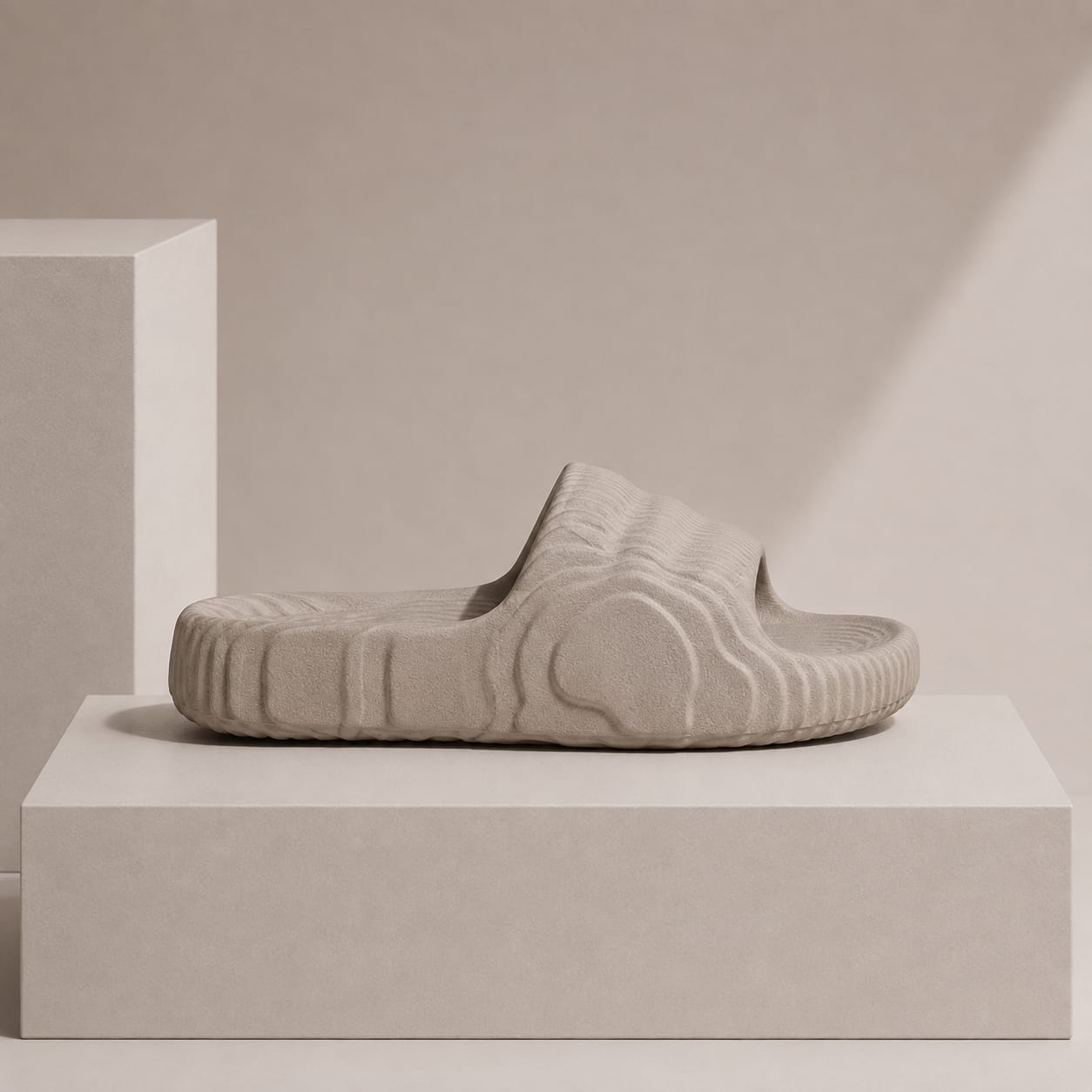 A single light beige slide sandal with a thick, textured wavy sole, displayed on a minimalist beige pedestal against a matching background.