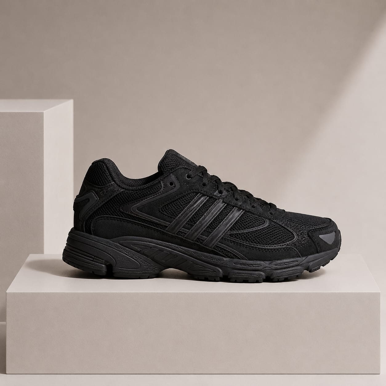A black athletic sneaker with three stripes, shown in profile on a minimalist beige platform against a neutral beige background.