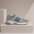A gray and white running shoe with navy blue stripes, displayed on a minimalist beige pedestal against a matching beige background.