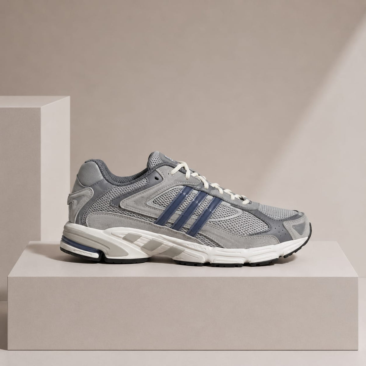 A gray and white running shoe with navy blue stripes, displayed on a minimalist beige pedestal against a matching beige background.