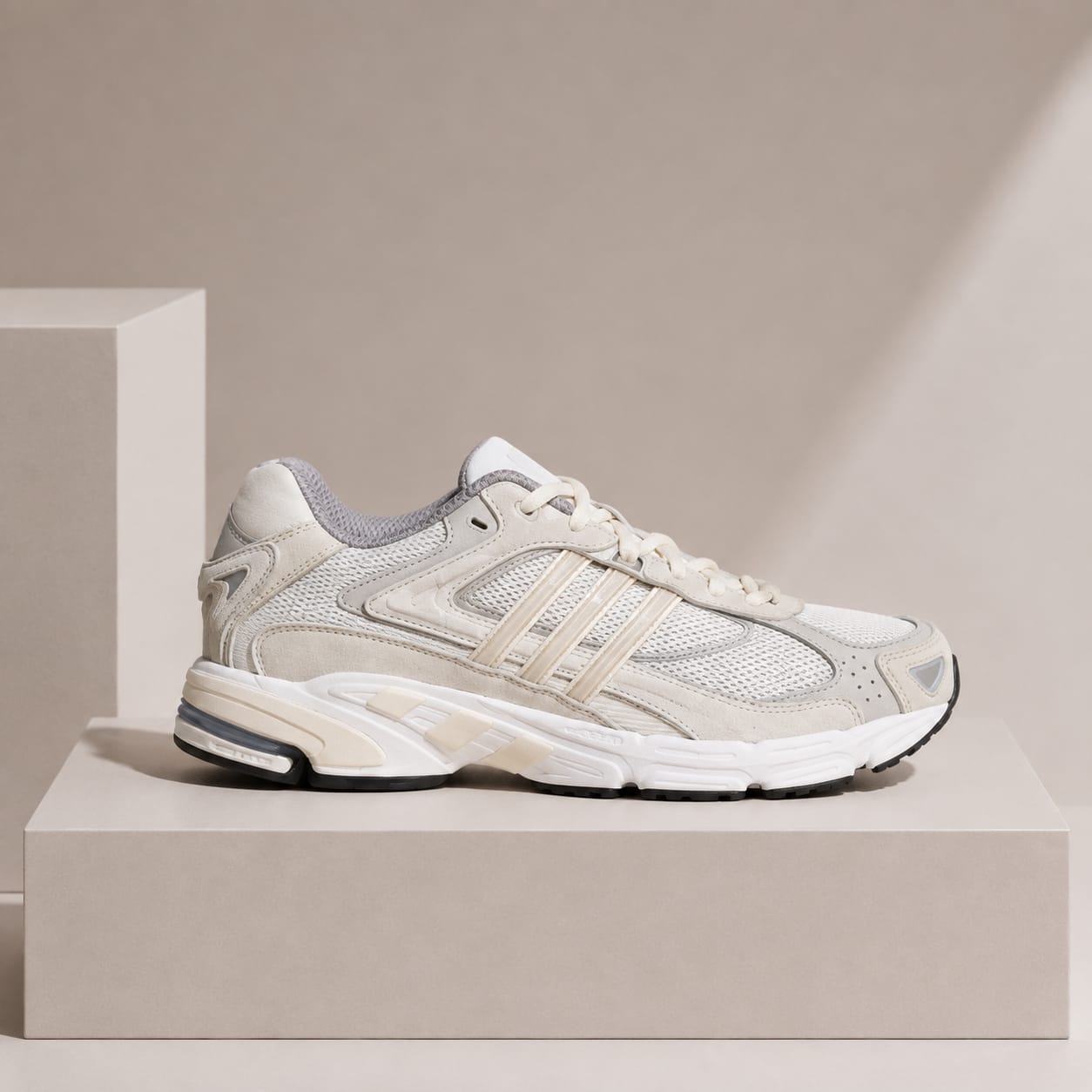 A single low-top athletic sneaker in off-white and beige mesh and suede, shown in side profile on a light beige pedestal against a beige studio background.