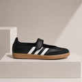 A black leather shoe with white three-stripes and a brown gum sole, displayed on a white geometric pedestal against a warm beige background.