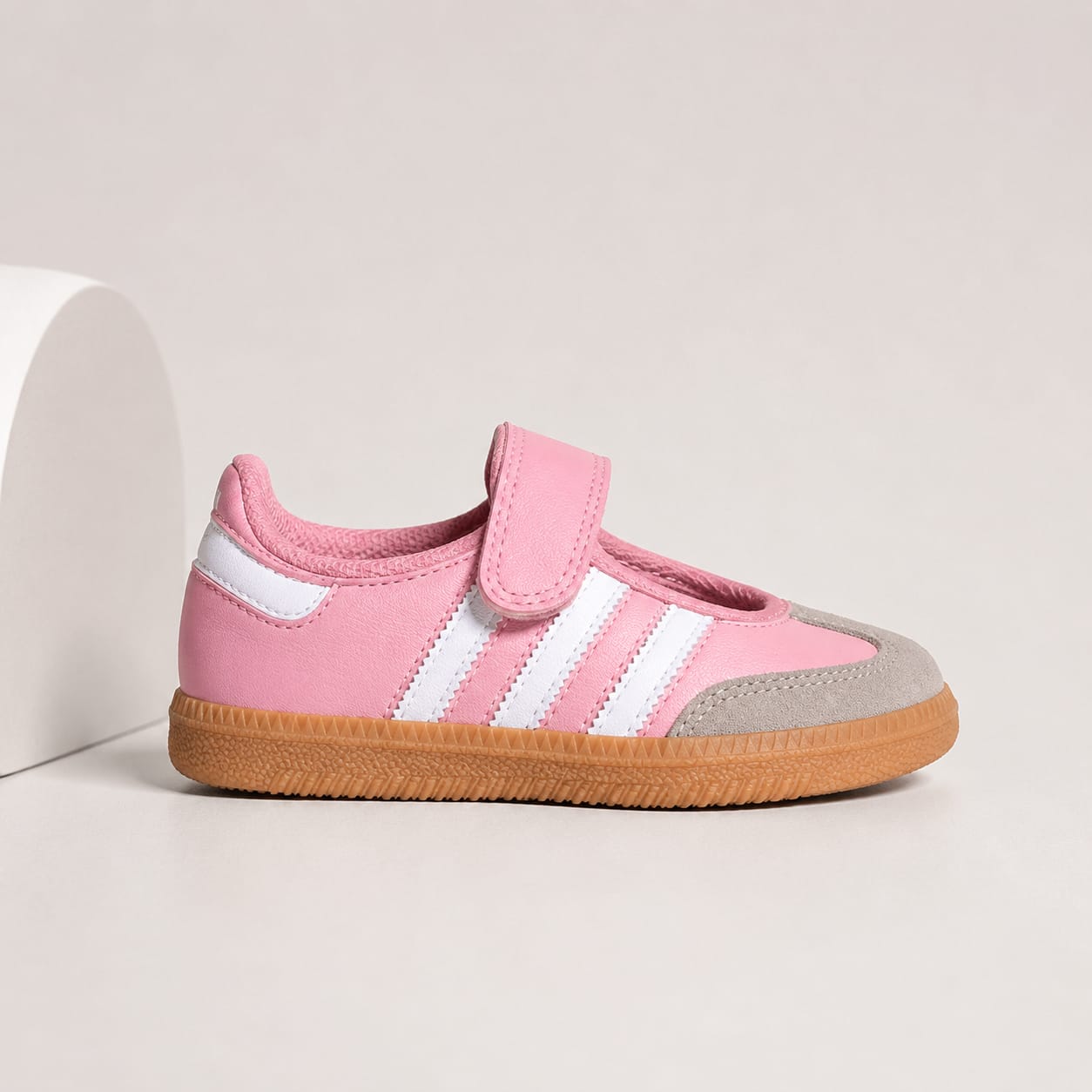 A pink toddler sneaker with white side stripes, a hook-and-loop strap, and a brown gum sole, shown on a light beige studio background.