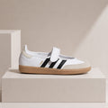 A white Adidas Gazelle sneaker with black three-stripes, a beige heel tab, and a gum sole, displayed on a minimalist beige pedestal.