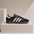 A black Adidas Samba sneaker with white stripes, a brown gum sole, and gold 'SAMBA' text, displayed on a beige pedestal against a beige background.
