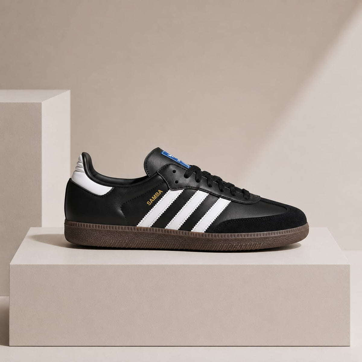 A black Adidas Samba sneaker with white stripes, a brown gum sole, and gold 'SAMBA' text, displayed on a beige pedestal against a beige background.
