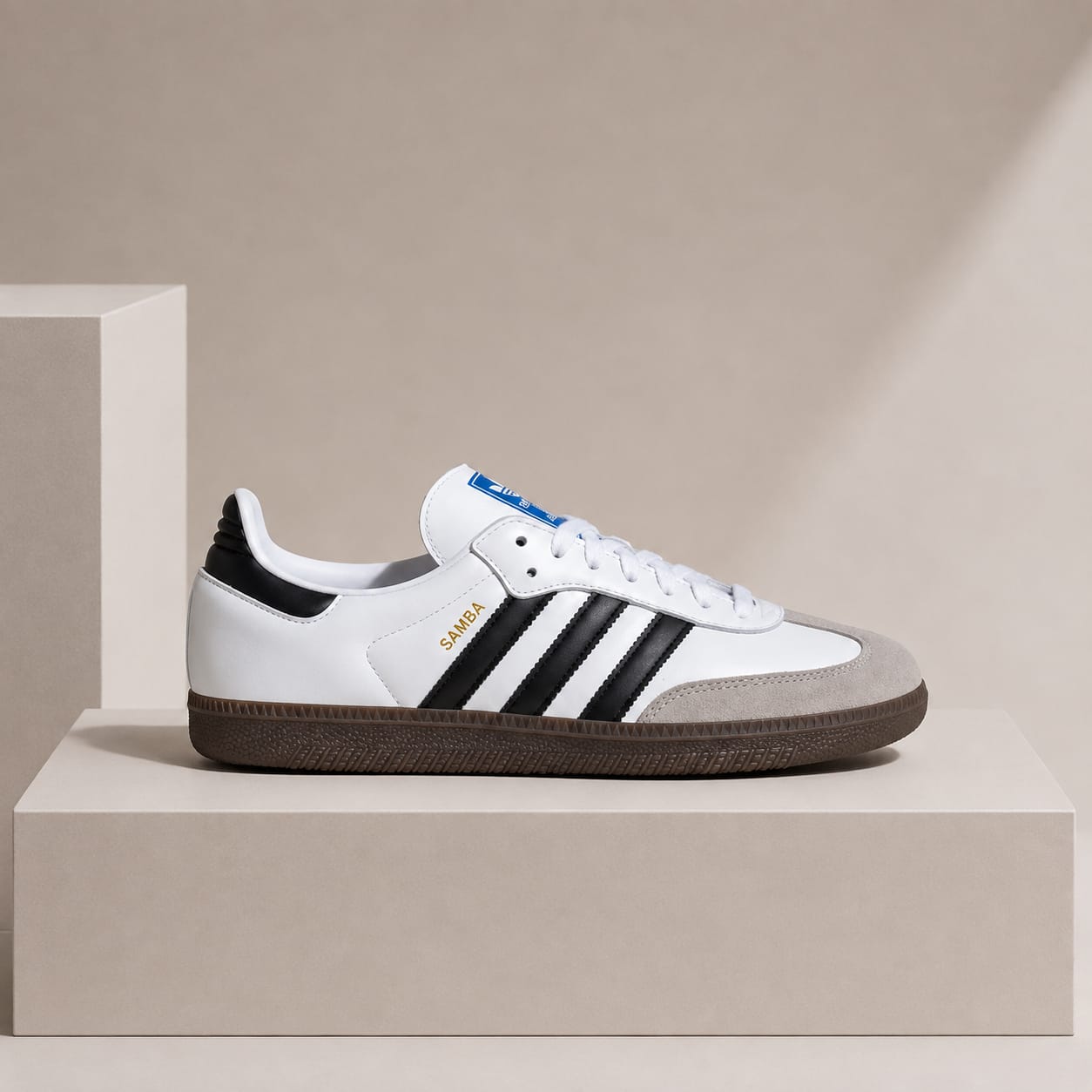 A white Adidas Samba sneaker with black three-stripes, a black heel tab, a brown gum sole, and gold 'SAMBA' text, displayed on a beige pedestal against a beige background.