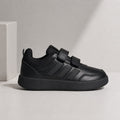 A black low-top sneaker with three hook-and-loop strap closures and three parallel side stripes, shown in profile against a light gray studio background with a white block prop.
