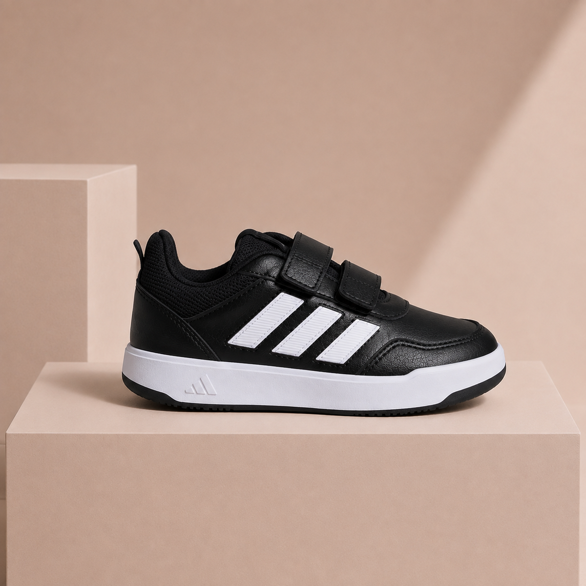 A black Adidas kids' sneaker with white three-stripes and a hook-and-loop strap closure, displayed on a beige geometric pedestal.