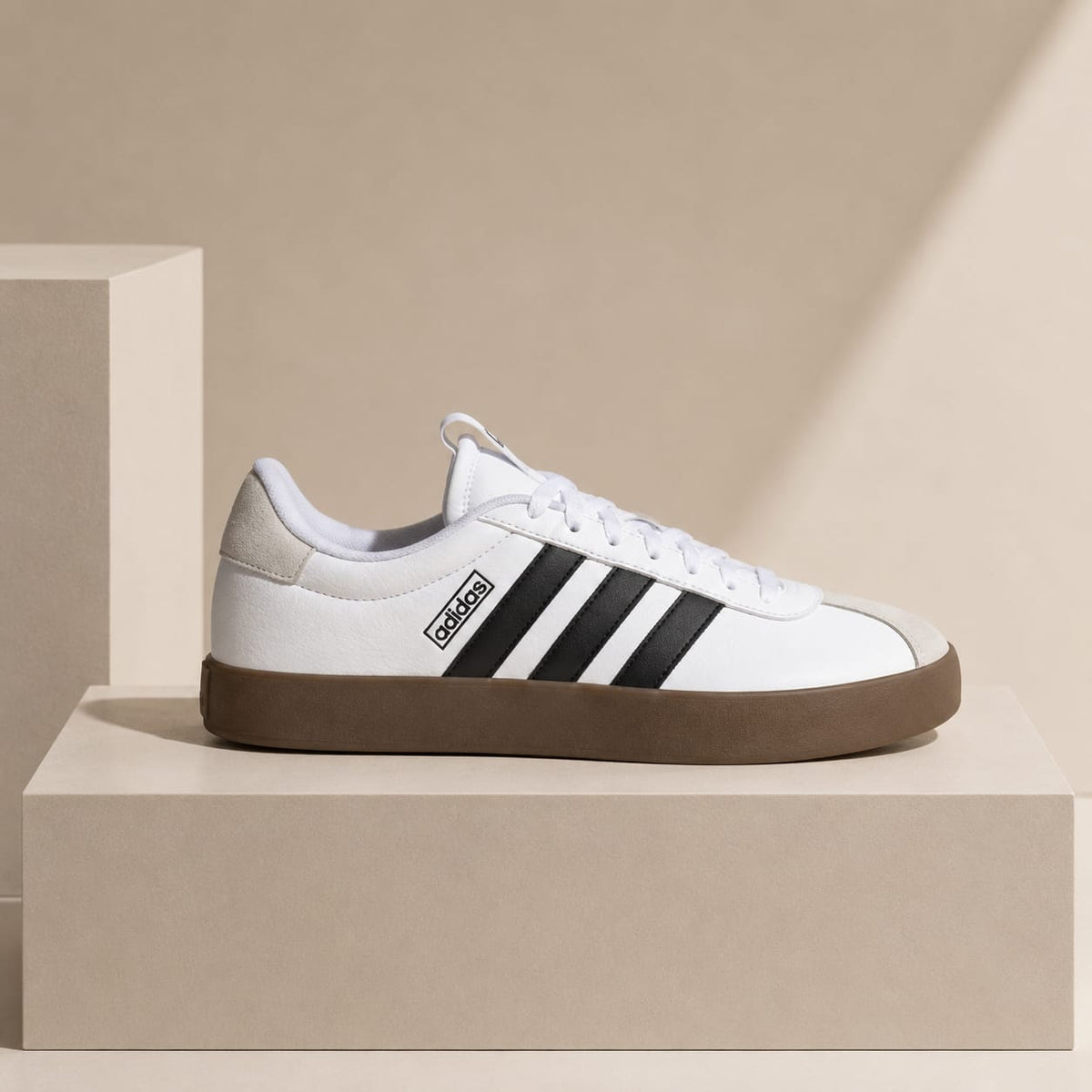 A white Adidas low-top sneaker with black three-stripes and a brown gum sole, displayed on a beige pedestal against a beige background.