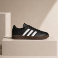 A black Adidas sneaker with three white stripes and a brown gum sole, displayed on a minimalist beige pedestal against a warm beige background.