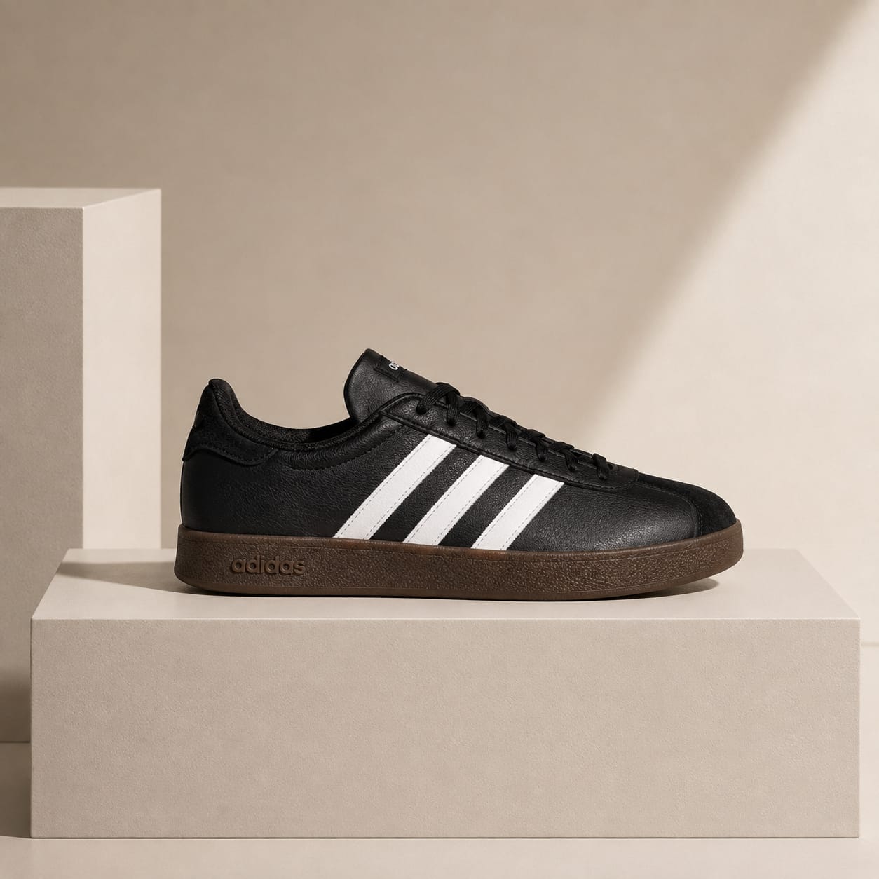 A black Adidas sneaker with three white stripes and a brown gum sole, displayed on a minimalist beige pedestal against a warm beige background.